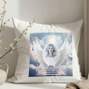 Puede incluir: Almohada blanca con una imagen impresa de una mujer con alas de &aacute;ngel y nubes. El texto dice "In Loving Memory Ashley Go Feb. 20, 1982 - Jan. 11, 2024".