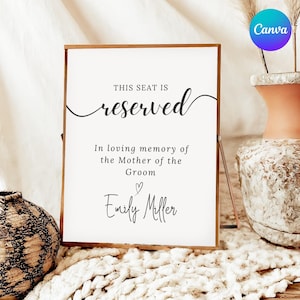 May include: A wooden framed sign with the text "THIS SEAT IS reserved" in elegant script. Below, it reads "In loving memory of the Mother of the Groom" and "Emily Miller" with a heart symbol. The sign is on a table with decorative vases.