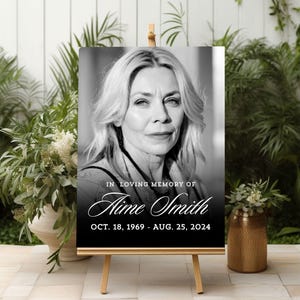 May include: A black and white memorial sign on a wooden easel. The sign features a portrait of a woman and the text "IN LOVING MEMORY OF Aime Smith OCT. 18, 1969 - AUG. 25, 2024."