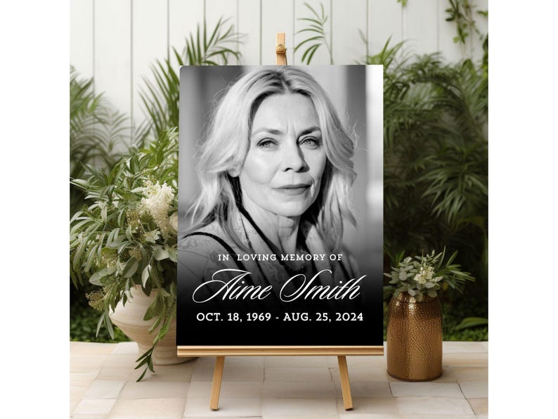 May include: A black and white memorial sign on a wooden easel. The sign features a portrait of a woman and the text "IN LOVING MEMORY OF Aime Smith OCT. 18, 1969 - AUG. 25, 2024."