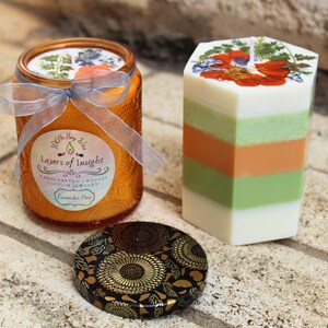 Pode incluir: Um conjunto de velas decorativas com uma vela de frasco de vidro laranja e uma vela de pilar em camadas. O frasco tem um rótulo com texto. A vela de pilar tem camadas de branco, verde e laranja, com decorações florais.