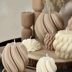 May include: A collection of decorative candles in various shapes and sizes. The candles are made of a beige and white wax and have a textured, ribbed design. The candles are arranged on a wooden surface with a brown tray.
