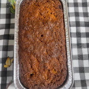 Puede incluir: Un pan recién horneado en una bandeja rectangular de papel de aluminio. El pan tiene una corteza dorada con una superficie ligeramente texturizada. El fondo es un patrón de cuadros blanco y negro.