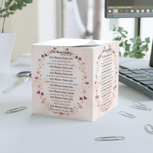 May include: A cube-shaped notepad featuring the text "The Beatitudes" and floral decorations. The text is arranged in a circular design, with the phrase "God Blesses those who" repeated. The notepad is placed on a white surface.