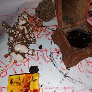 Könnte beinhalten: Eine Anordnung von Objekten, darunter eine kleine Cherub-Statue, eine Holzkiste, ein Tannenzapfen und ein gelbes rechteckiges Objekt mit Nadeln und dunkler roter Flüssigkeit. Die Objekte liegen auf einer weißen Oberfläche mit roten und schwarzen Markierungen.