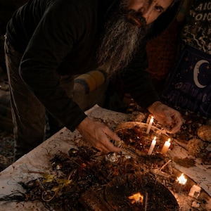Pode incluir: Uma pessoa com um moletom preto com capuz se inclina sobre uma mesa com velas e objetos rituais. A mesa é coberta com um material escuro e orgânico, e um prato de madeira com chamas está no centro.