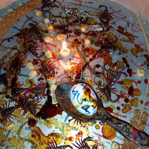Op de afbeelding: Een rituele scène met een metalen dienblad met talrijke schorpioenen, kleine witte voorwerpen en een brandende kaars. Een versierd ei rust in een houten lepel. De lade is bedekt met een roodbruine vloeistof en bezaaid met zaden.