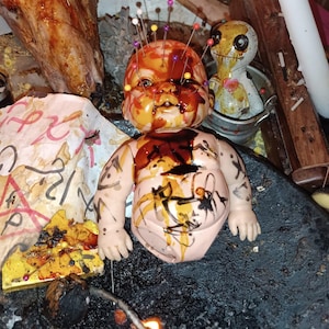 Pode incluir: Uma boneca com alfinetes na cabeça e manchas escuras no corpo está sentada em uma superfície escura. Uma boneca pequena está em um balde atrás dela. Uma vela acesa e outros itens rituais estão ao fundo.