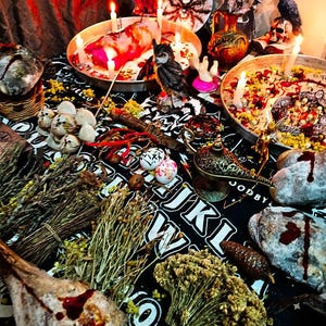 Könnte beinhalten: Ein schwarz-weißes Ouija-Brett mit verschiedenen Gegenständen darauf, darunter Kerzen, getrocknete Kräuter, ein Schädel, eine Keramik-Eule und eine kleine goldene Lampe. Die Gegenstände sind in einem Kreis um das Brett angeordnet, das auf einem schwarzen Tuch mit weißem Muster liegt.