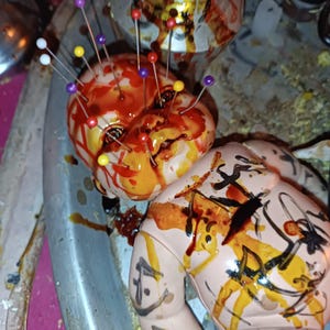 May include: A plastic doll head and torso featuring multiple colourful sewing pins inserted into the scalp and face. The doll is covered in splatters of red and yellow paint, with black hand-drawn markings across the chest and arms.