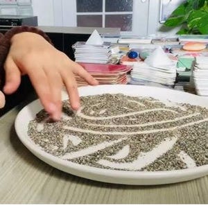 May include: A white ceramic plate with a design created by drawing lines in a layer of dark grey sand.