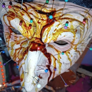 Op de afbeelding: Een grotesk masker met een bleke teint, bevlekt met bruine en rode tinten en versierd met talrijke spelden in verschillende kleuren. Het masker heeft een sinistere uitstraling, met een prominente neus en oogkassen.