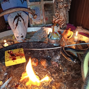 May include: A dimly lit scene featuring a variety of objects. A burning flame is in the foreground, with a snake, a doll, and a statue of an angel in the background. Other items include a skull, candles, and a small wooden block.