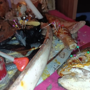 Puede incluir: Una disposición de objetos rituales, incluyendo un objeto negro, partes de animales y una rana, adornados con alfileres. Una vela blanca y un aplicador con punta roja también están presentes, sobre una tela rosa.