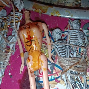 Puede incluir: Una muñeca con un torso pintado y un diseño de cruz roja, rodeada de decoraciones de esqueleto y cuerda. La muñeca está cubierta con una sustancia amarilla, y la escena se desarrolla sobre una superficie rosa.