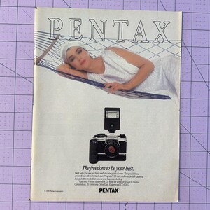 May include: Vintage advertisement for Pentax cameras. The ad features a woman reclining on a hammock, with a Pentax camera in the foreground. The text reads "The freedom to be your best."