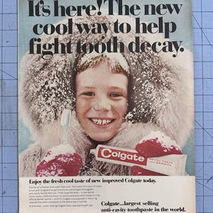 May include: Vintage advertisement for Colgate toothpaste. The ad features a child wearing a fur-lined hood and mittens, holding a toothbrush and a tube of Colgate. Text reads, "It's here! The new cool way to help fight tooth decay."