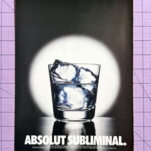 May include: A clear glass filled with ice cubes and liquid, set against a dark background with a bright circular light. The text "ABSOLUT SUBLIMINAL" is displayed in white at the bottom of the image.