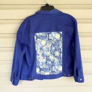 May include: A blue denim jacket with a vibrant, artistic design on the back. The design features a swirling pattern of blues, whites, and yellows, reminiscent of a starry night. The jacket has button closures and pockets.