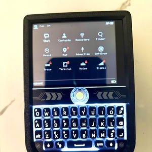 May include: A black handheld device with a screen displaying icons and text. The device has a backlit keyboard with letters and numbers. The screen shows icons for chat, contacts, and settings. The device has the brand name "LILYGO®" on the bottom.