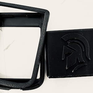 Puede incluir: Funda de teléfono negra impresa en 3D y un emblema cuadrado a juego. La funda del teléfono tiene un marco rectangular con una superficie texturizada. El emblema presenta un diseño de casco espartano.