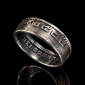 Könnte beinhalten: Ein silberfarbener Ring mit einem komplizierten Muster aus geometrischen Formen und Symbolen. Der Ring hat eine strukturierte Oberfläche und ist vor einem schwarzen Hintergrund platziert, mit einer Reflexion darunter.