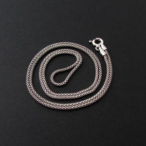 May include: A silver-colored necklace with a textured, woven design. The necklace is laid out on a black surface, with a clasp at one end. The chain is approximately 18 inches long.