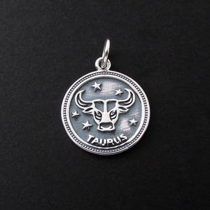 May include: A silver-colored Taurus zodiac pendant. The pendant features a bull's head in the center, surrounded by stars and the word "TAURUS". The pendant has a loop at the top for attaching to a chain.