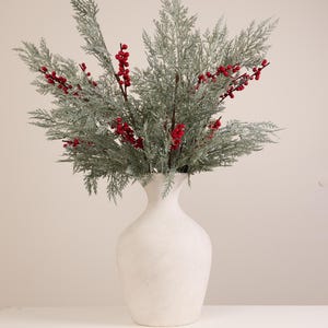Könnte beinhalten: Eine weiße Vase mit einem dekorativen Arrangement aus bereiften grünen Zweigen und leuchtend roten Beeren. Die Vase hat eine strukturierte Oberfläche und eine klassische Form, geeignet für die Inneneinrichtung.