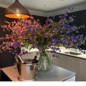 Peut inclure: Un grand bouquet de fleurs violettes et roses dans un vase en verre transparent. Un seau à glace argenté avec des bouteilles est placé à gauche. Une lampe suspendue en métal sombre est au-dessus.
