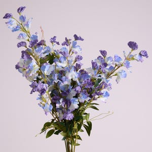 May include: A bouquet of artificial flowers with a variety of blue and purple blooms. The flowers have a delicate, layered petal design and are accented by green leaves and stems. The arrangement is set against a soft, neutral background.