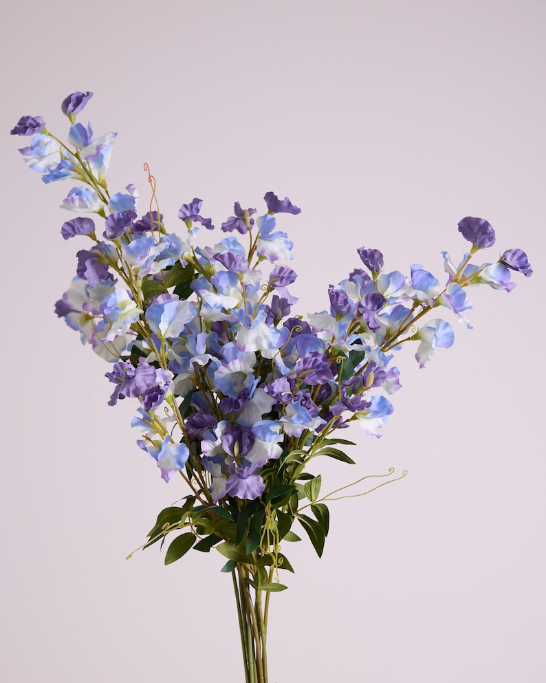 May include: A bouquet of artificial flowers with a variety of blue and purple blooms. The flowers have a delicate, layered petal design and are accented by green leaves and stems. The arrangement is set against a soft, neutral background.