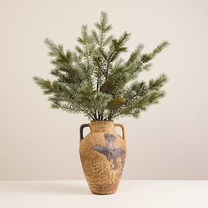 May include: A decorative arrangement featuring a faux evergreen plant in a textured, brown ceramic vase. The vase has a unique, aged appearance with dark brown accents. The plant has green and blue-green needles.