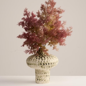 May include: A decorative vase with a wide, rounded top and a textured, speckled pattern in cream and green. The vase holds a cluster of pink and yellow-green faux foliage, creating a striking floral arrangement. The vase is approximately 10 inches tall.