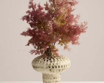 May include: A decorative vase with a wide, rounded top and a textured, speckled pattern in cream and green. The vase holds a cluster of pink and yellow-green faux foliage, creating a striking floral arrangement. The vase is approximately 10 inches tall.