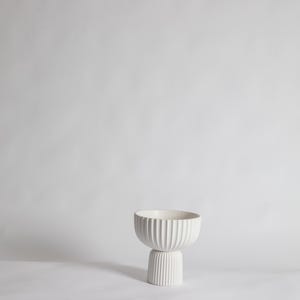 May include: A white, ribbed ceramic bowl with a pedestal base. The bowl has a rounded shape with vertical grooves, and the base is a shorter, cylindrical form with matching grooves. The piece is set against a plain white background.