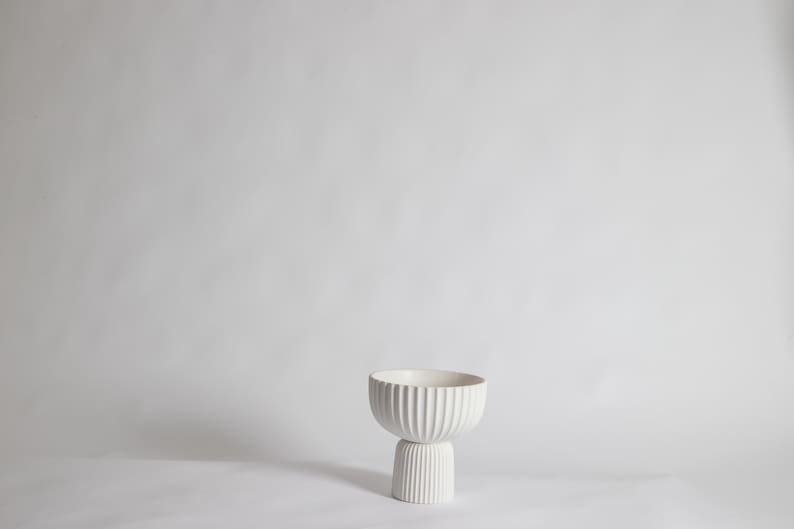 May include: A white, ribbed ceramic bowl with a pedestal base. The bowl has a rounded shape with vertical grooves, and the base is a shorter, cylindrical form with matching grooves. The piece is set against a plain white background.