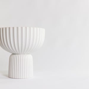 May include: A white, modern ceramic bowl with a ribbed texture, resting on a matching pedestal base. The bowl has a wide, rounded shape, and the base is cylindrical. The design is minimalist and elegant, suitable for home decor.