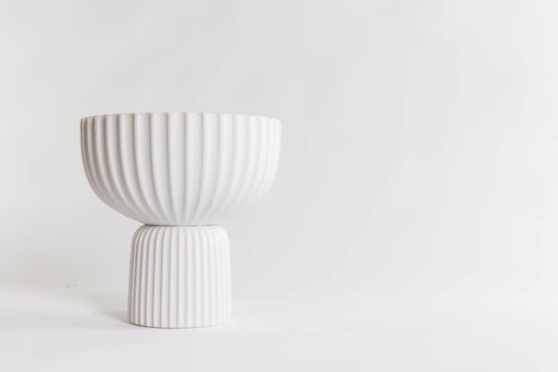 May include: A white, modern ceramic bowl with a ribbed texture, resting on a matching pedestal base. The bowl has a wide, rounded shape, and the base is cylindrical. The design is minimalist and elegant, suitable for home decor.