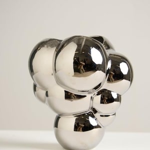 May include: A modern, silver-colored sculpture with a cluster of spherical shapes. The reflective surface of the spheres creates a mirrored effect, capturing the surrounding environment. The sculpture is a decorative art piece.