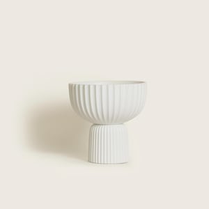May include: A white ceramic bowl with a ribbed exterior, resting on a smaller, matching ribbed base. The bowl has a wide, open top and a rounded shape. The design is minimalist and modern, suitable for home decor.