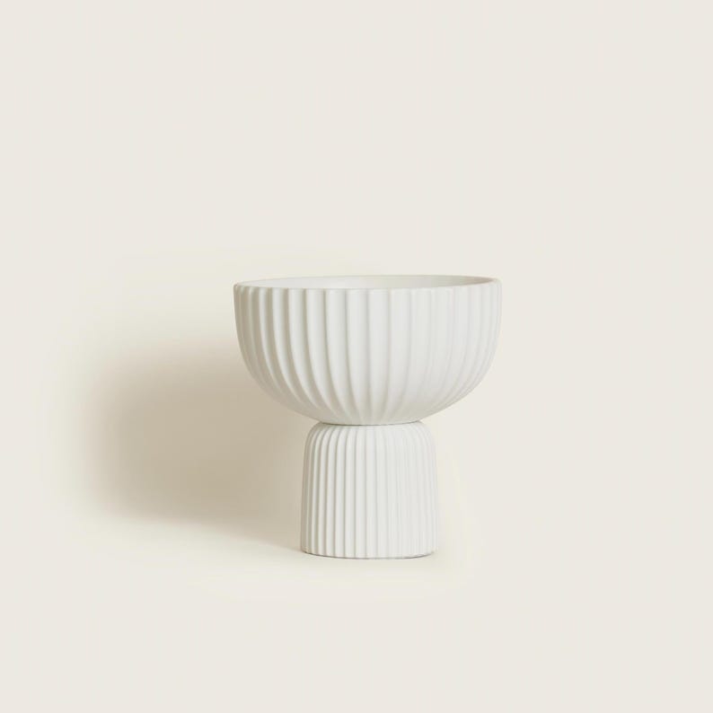 May include: A white ceramic bowl with a ribbed exterior, resting on a smaller, matching ribbed base. The bowl has a wide, open top and a rounded shape. The design is minimalist and modern, suitable for home decor.