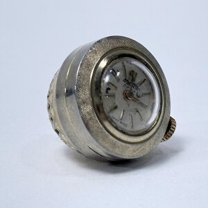 Op de afbeelding: Een vintage zilverkleurige ringhorloge met een ronde wijzerplaat. De wijzerplaat heeft een lichtgekleurde achtergrond met zwarte cijfers en wijzers. Het horloge is gemarkeerd met "Swiss Made". De ring heeft een textuur.