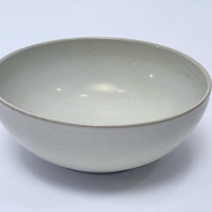 May include: A pale grey ceramic bowl with a rounded form. The bowl features a smooth interior and a subtly darker rim. It is set against a white backdrop, emphasising its simple design and neutral colour.