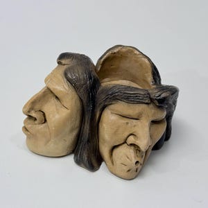 May include: A beige sculpture depicting three faces with closed eyes and dark hair. The sculpture is carved from a single piece of material, with a rough texture and visible details. The faces are stylized.