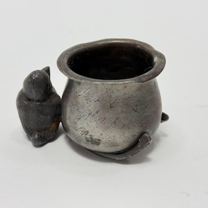 Op de afbeelding: Een zilveren tinnen beker met een vogelbeeldje aan de zijkant. De beker heeft een inscriptie met de tekst "Our Baby Mother 1916".