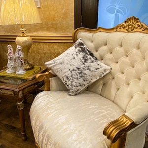 Op de afbeelding: Een gecapitonneerde crèmekleurige fauteuil met gouden accenten en een decoratief kussen. Het kussen heeft een koeienhuidpatroon in zwart en wit. Links een tafel met een lamp en beeldjes.