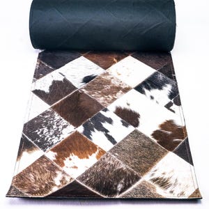 May include: A brown and white cowhide rug with a diamond pattern. The rug is rolled up and has a black backing.