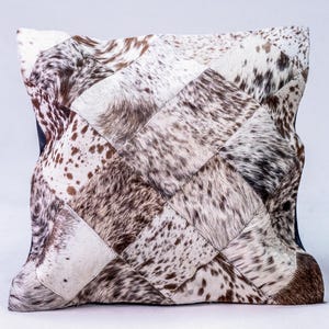 May include: A square pillow with a patchwork design made from cowhide. The pillow is made of different pieces of cowhide, each with a different pattern of brown and white spots. The pillow is a light brown color overall.
