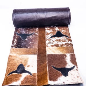 May include: A brown leather-backed runner rug with a patchwork design of cowhide. The rug has four squares with black leather cow head silhouettes in each square.